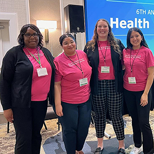 Four female College of Social Work students pose in front of a display at the Advancing Health Equity in South Carolina summit.