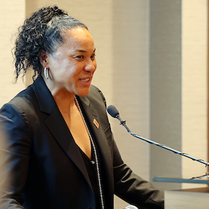 woman with black hair in a pony tail and a black suit on speaks at a lectern