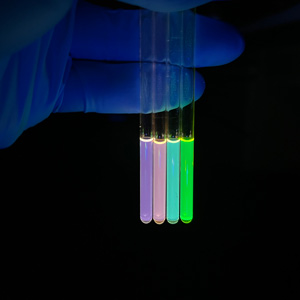 A gloved hand holds four tubes displaying fluorescent nanocyrstals in a dark space.