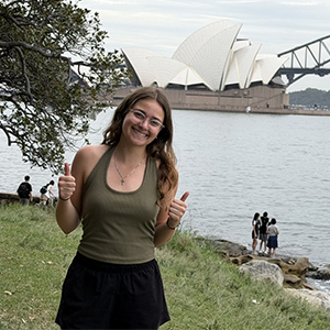 Ashley Chittum smiles and gives two thumbs up in Australia, with the Sydney Opera House behind her