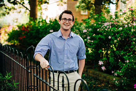 male student wearing glasses in blue check shirt standing behind an iron fence