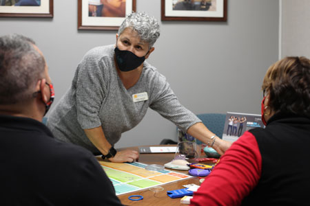 Woman with gray hair, gray shirt and black mask stands in front of a table with health items. Man and woman at table with backs to viewer.