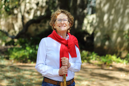 woman wearing glasses in white shirt with red sweater tied around shoulders and holding a walking stick with foliage in the background