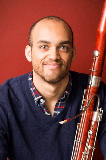 male musician smiling holding a bassoon wearing a blue sweater and red background