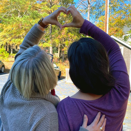 two women facing away from the camera embrace and make a heart symbol with their hands