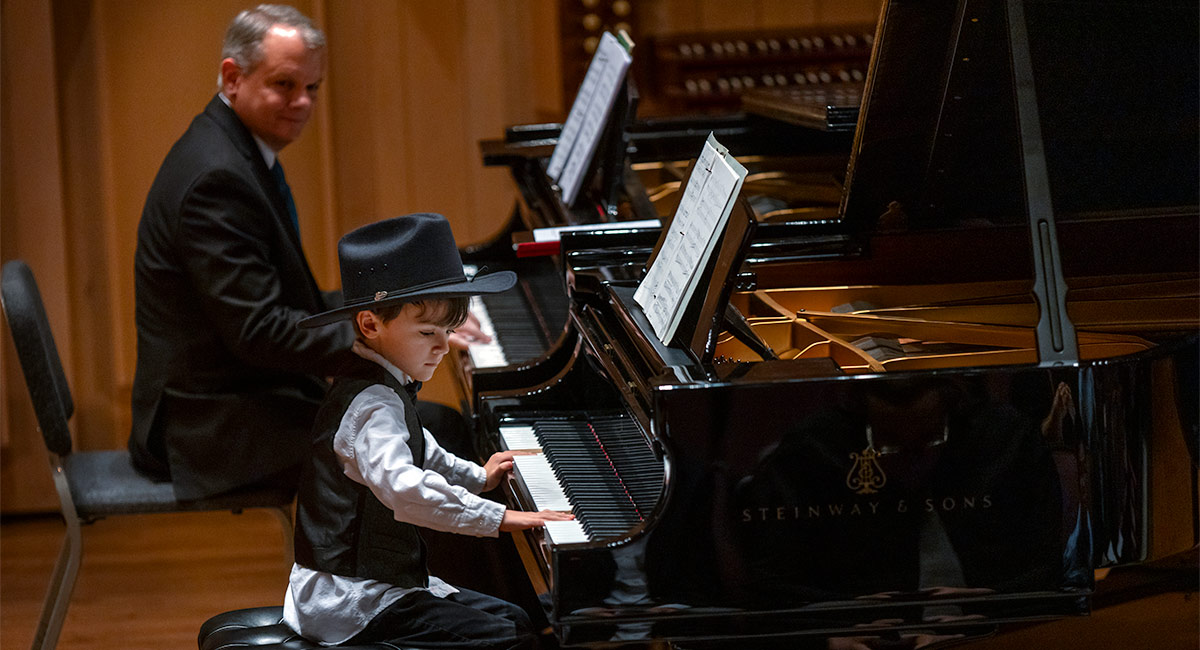 Boy in cowboy hat playing piano as teacher looks on