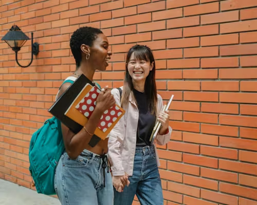 Two schoolgirls of African American and Asian descent, respectively, stroll alongside a brick wall.